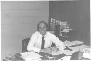A black and white photo of a man at his desk with papers strewn on the surface. He is wearing a white dress shirt and tie and is smiling at the camera. 