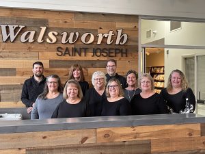 A current photo of the St. Joseph Walsworth office. A group of 10 individuals are smiling and posing in front of the "Walsworth Saint Joseph" sign inside the office. 