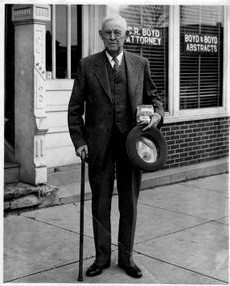 A photo of Ovid Bell as an older man. He is wearing a suit with a cane and a hat in his hand. He is standing in front of Boyd & Boyd Abstracts