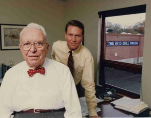 John Bell with a white collared shirt and red bowtie with John O Bell leaning behind him in an office. 