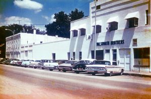 The exterior of the Walsworth headquarters in what looks to be the 1960s. Several vintage cars are parked in front of a white building with a sign reading "Walsworth Brothers" on it.
