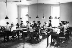 Eleven women, in what appears to be the 1940s, sit in rows while they work on typewriters