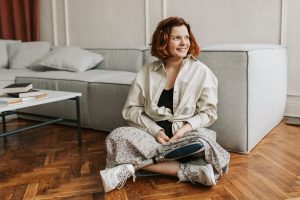 A photo of a smiling woman with a prosthetic leg. She is sitting on the floor leaning against a grey couch looking away from the camera.