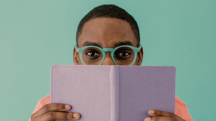 A man with green glasses holding a book in front of his face.