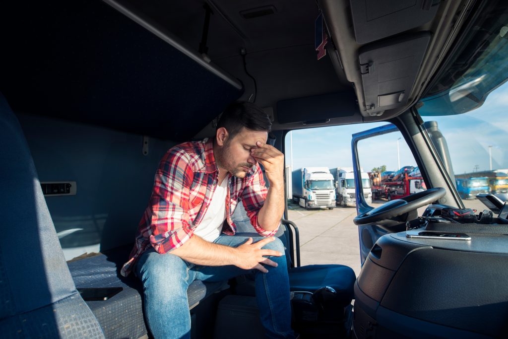 A truck driver in the trucking industry sitting in the front seat with his hands on his face, in frustration.