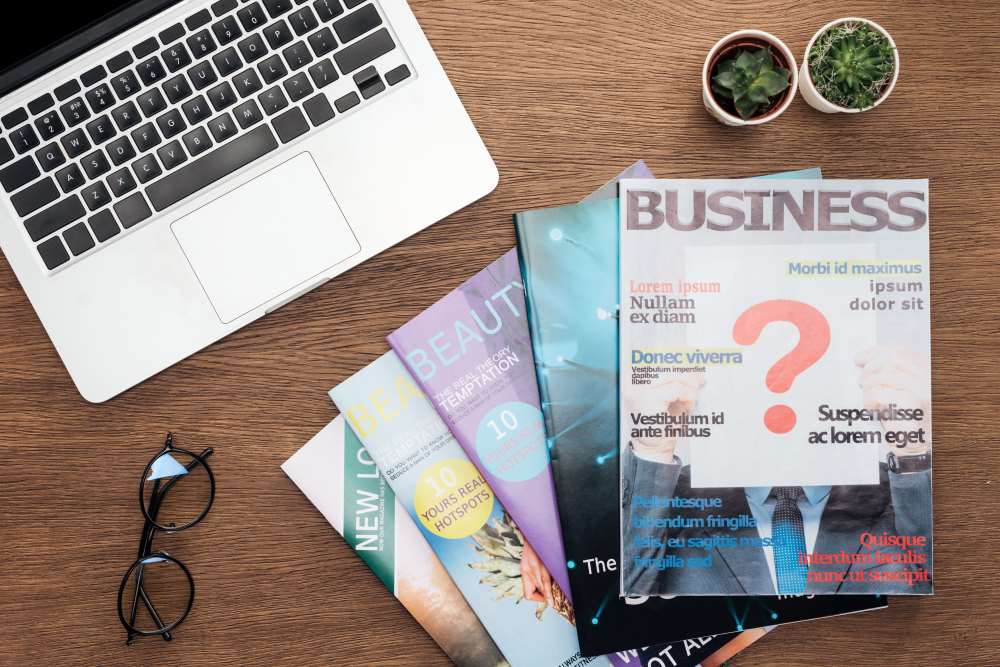 A stack of magazines on a desk next to a laptop and a pair of glasses. The top magazine has a question mark on the front.