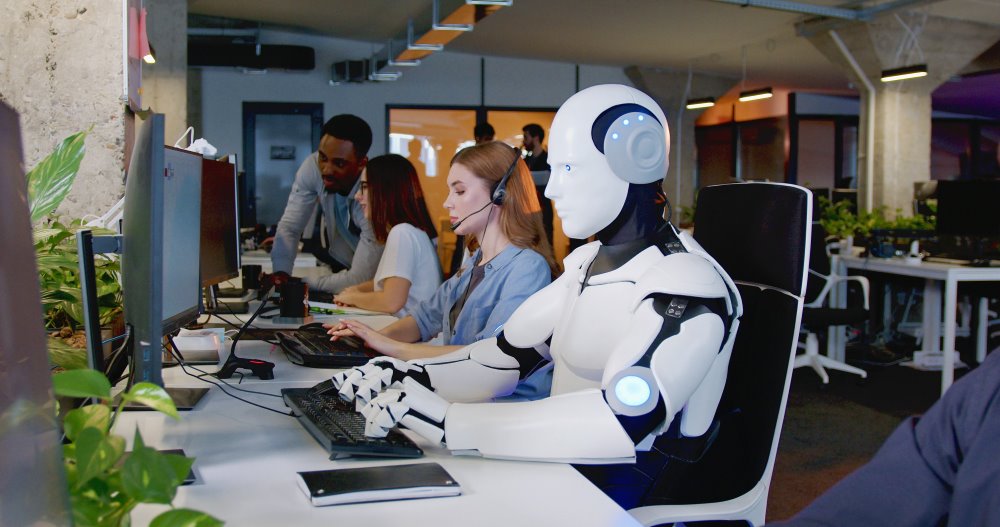 A white robot working at a computer next to his human coworkers