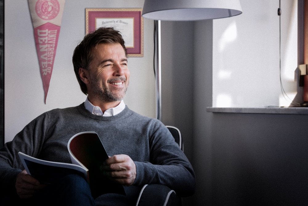 A smiling man reading a magazine with his diploma and a University of Denver banner on the wall behind him